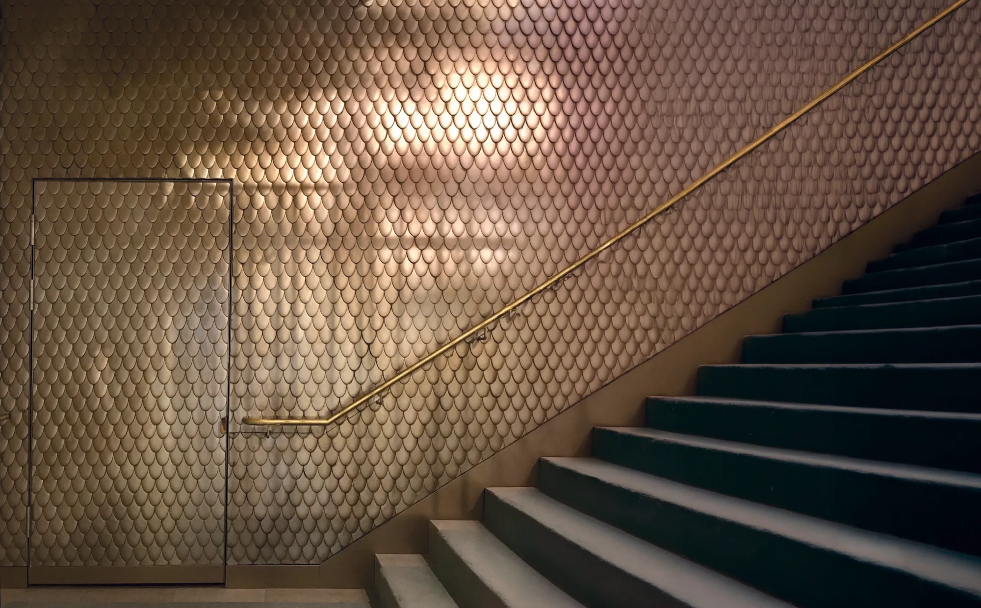 Glastür eingelassen in eine Wand mit bronzener Schuppen-Struktur, daneben Treppe mit goldenem Handlauf