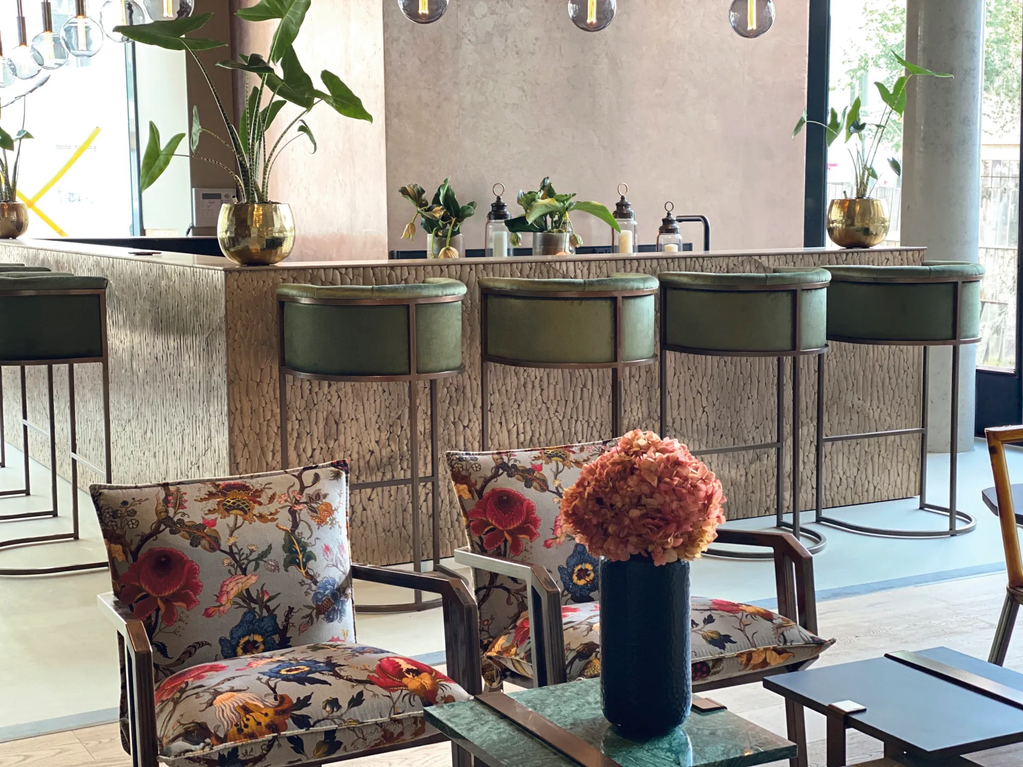 Hotelbar mit grünen Lederhockern, floral bezogenen Sesseln und Tresen in haptischer Naturstein-Optik vor Glasfront