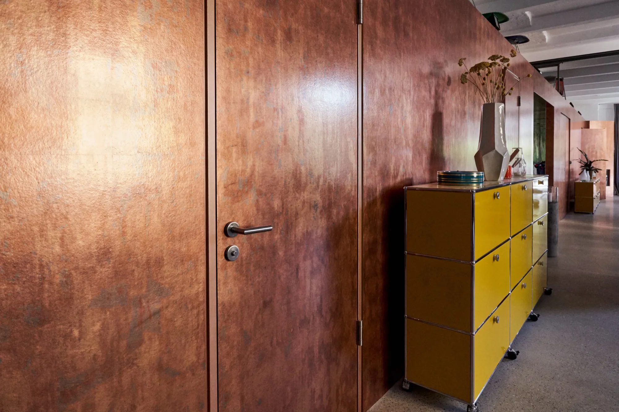 Innenraum mit Wand und Türen in Blattkupfer mit warmer Patina; gelbes Sideboard im Vordergrund